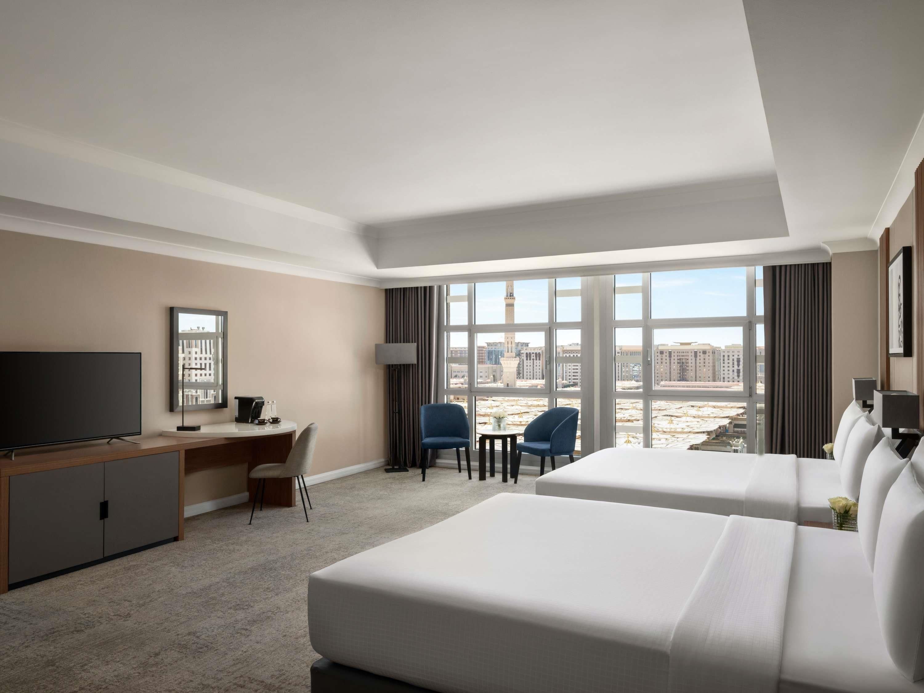 A hotel room with two beds, a desk, a TV, and a view of a cityscape through large windows.
