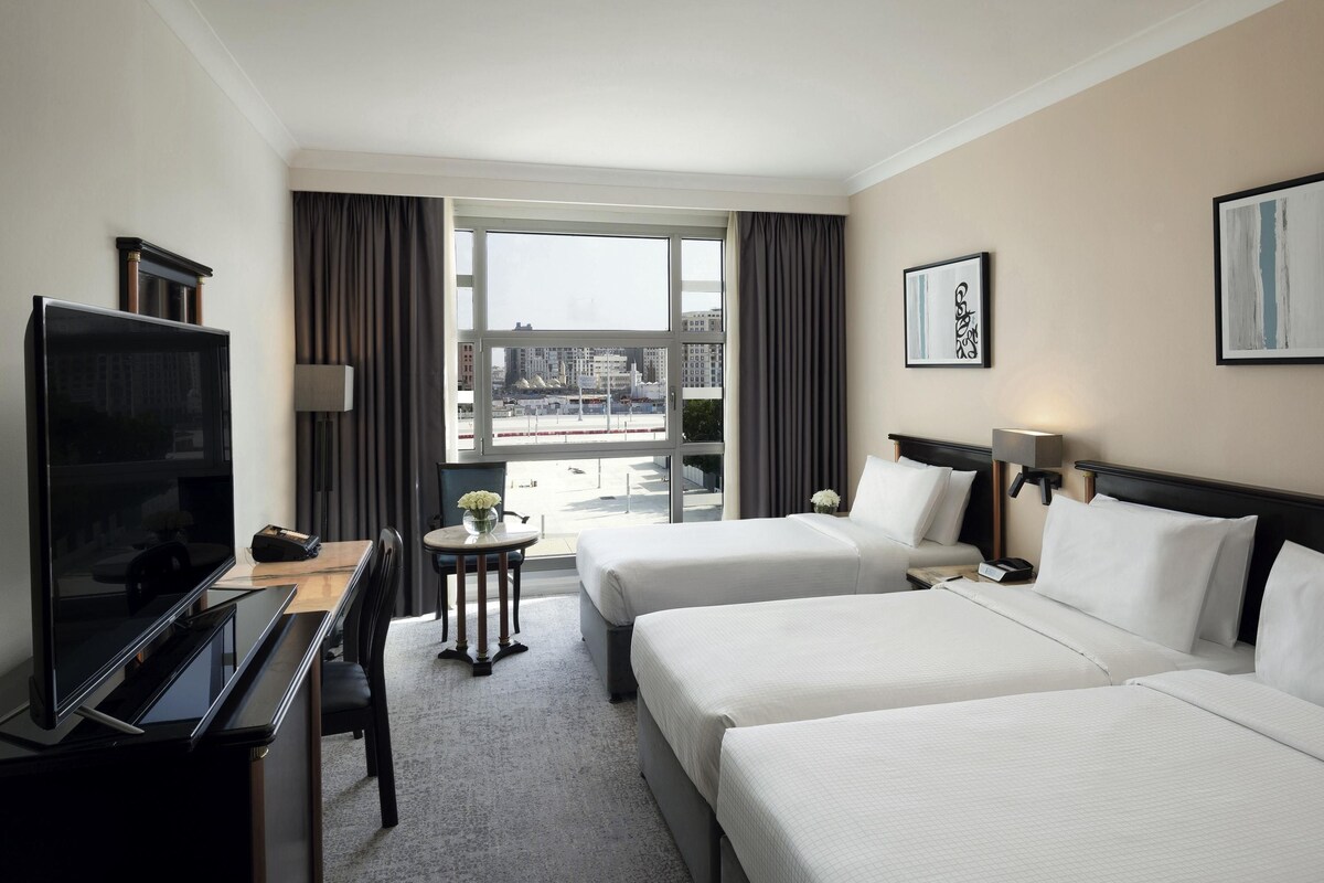 Habitación de hotel con dos camas, un escritorio con televisor, una mesita con un florero y vistas a la ciudad a través de amplios ventanales.