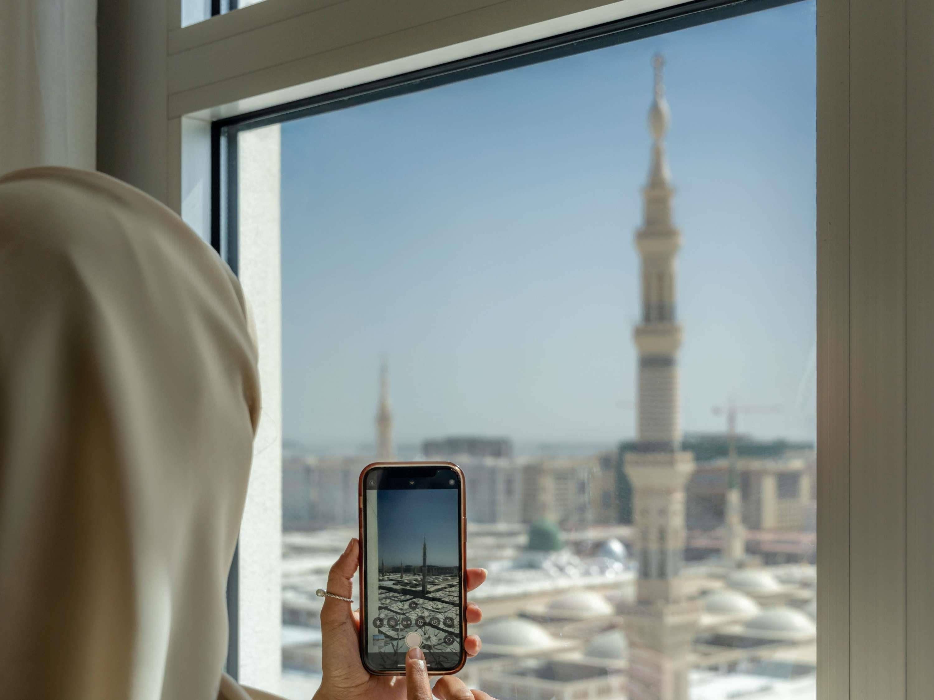 A person holding a smartphone with a view of a mosque's minaret through a window.
