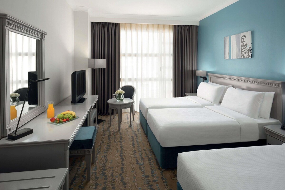 Habitación de hotel con dos camas, un escritorio con computadora, una silla, una mesita con un florero y un cuadro en la pared.