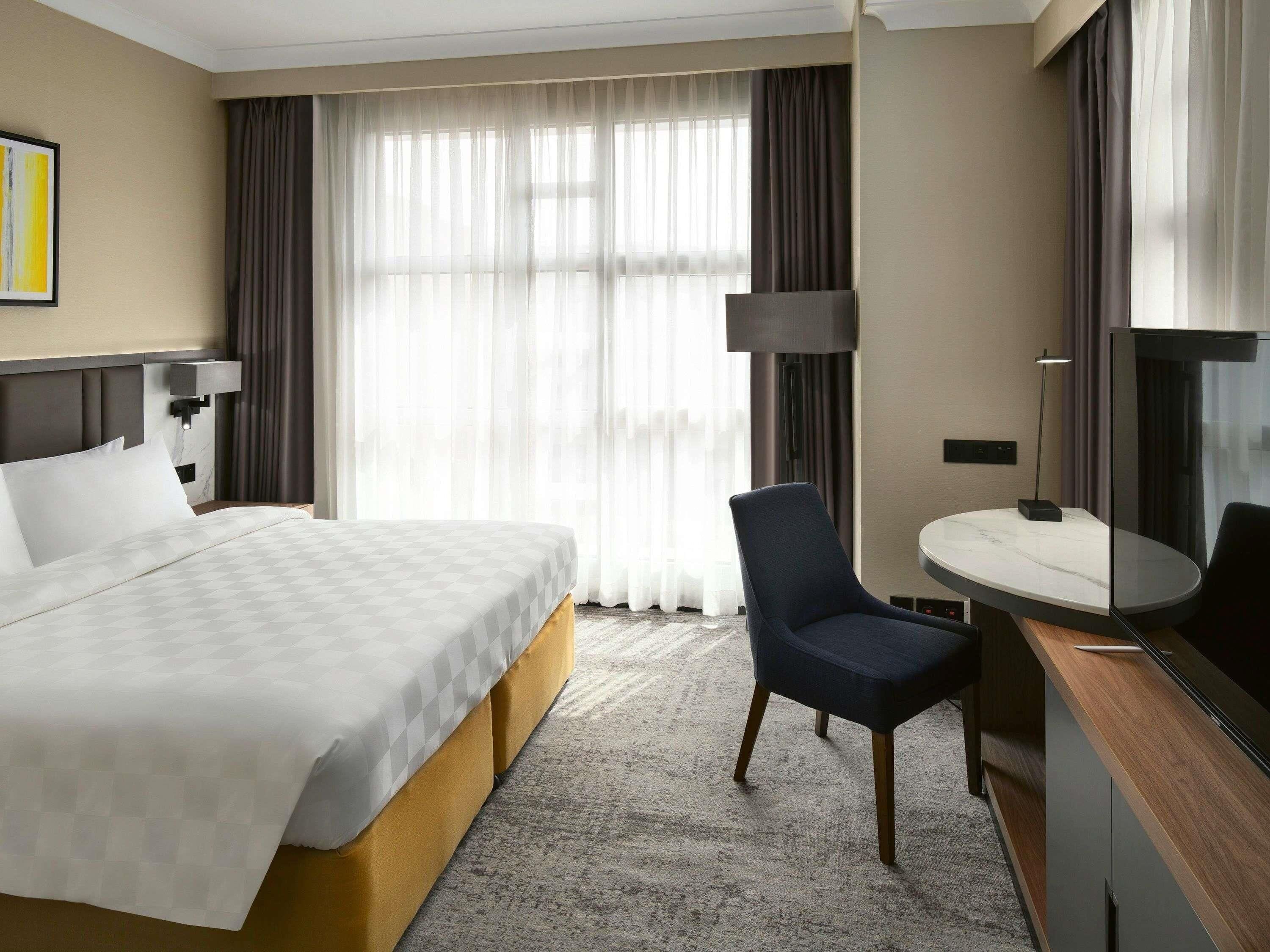 Habitación de hotel con una cama grande, un escritorio con silla, una lámpara y un ventanal con cortinas.