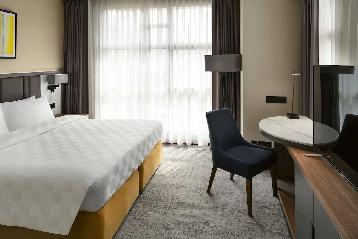 Habitación de hotel con una cama grande, un escritorio con silla, una lámpara y un ventanal con cortinas.