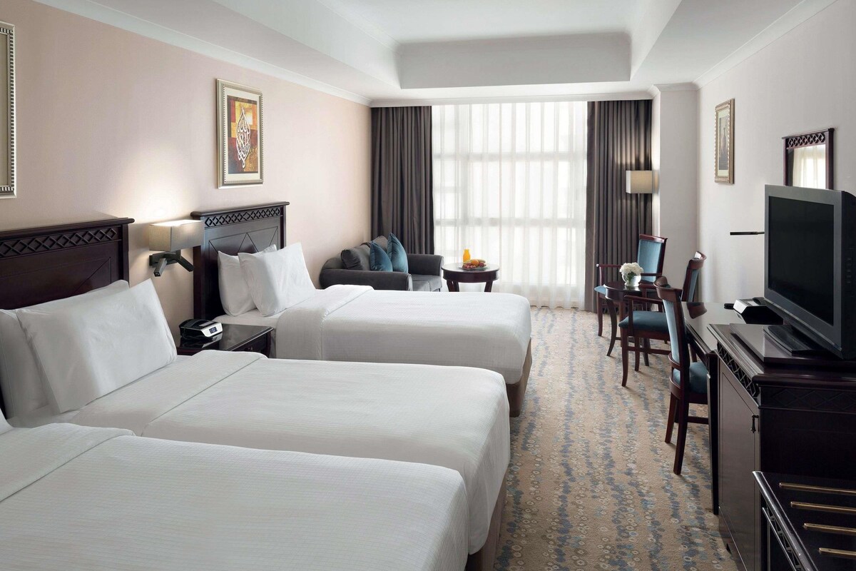 Habitación de hotel con dos camas, televisor, escritorio con silla y una ventana grande con cortinas.