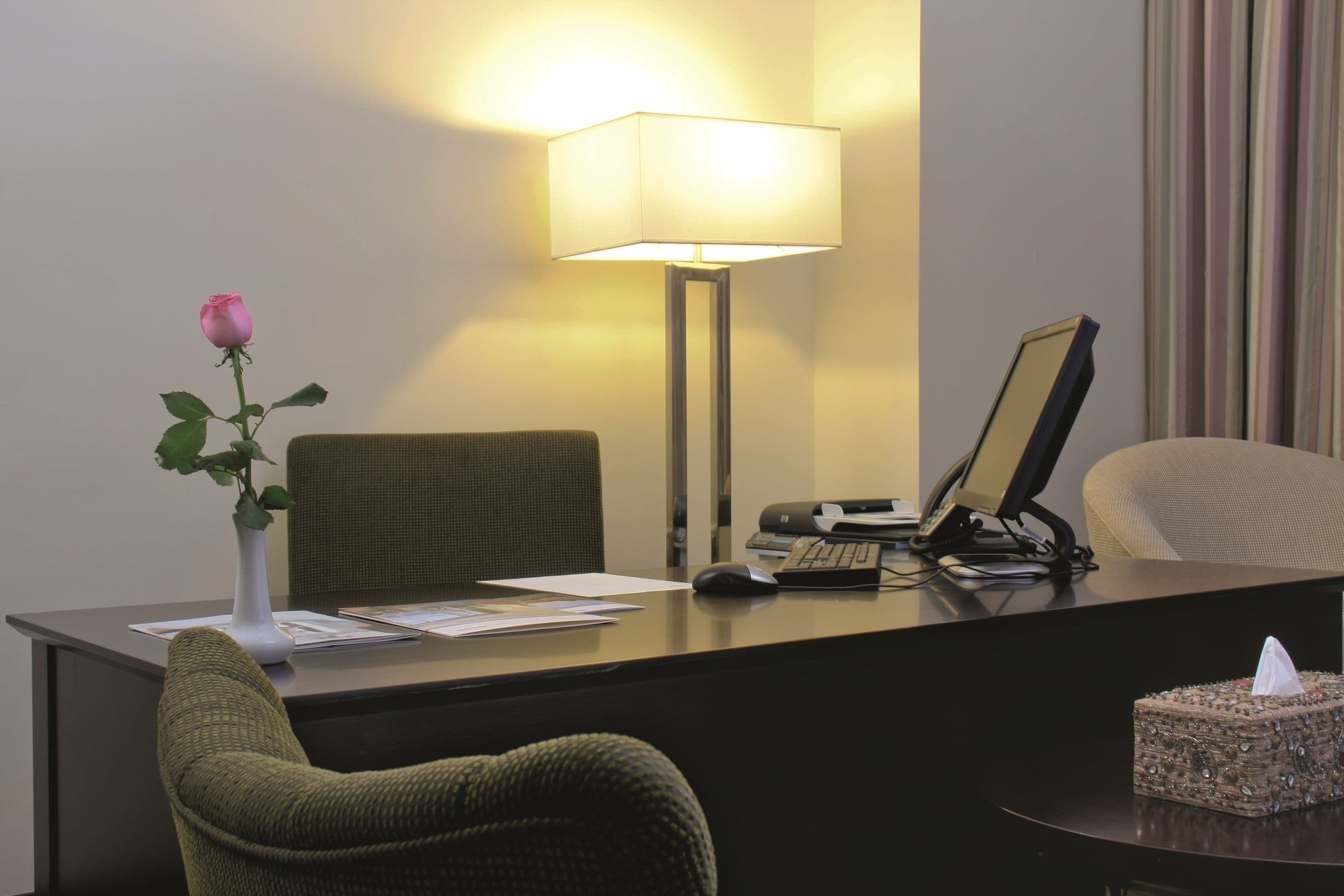 Un mostrador de recepción de hotel con un jarrón de flores, una lámpara de escritorio, una computadora y una silla.