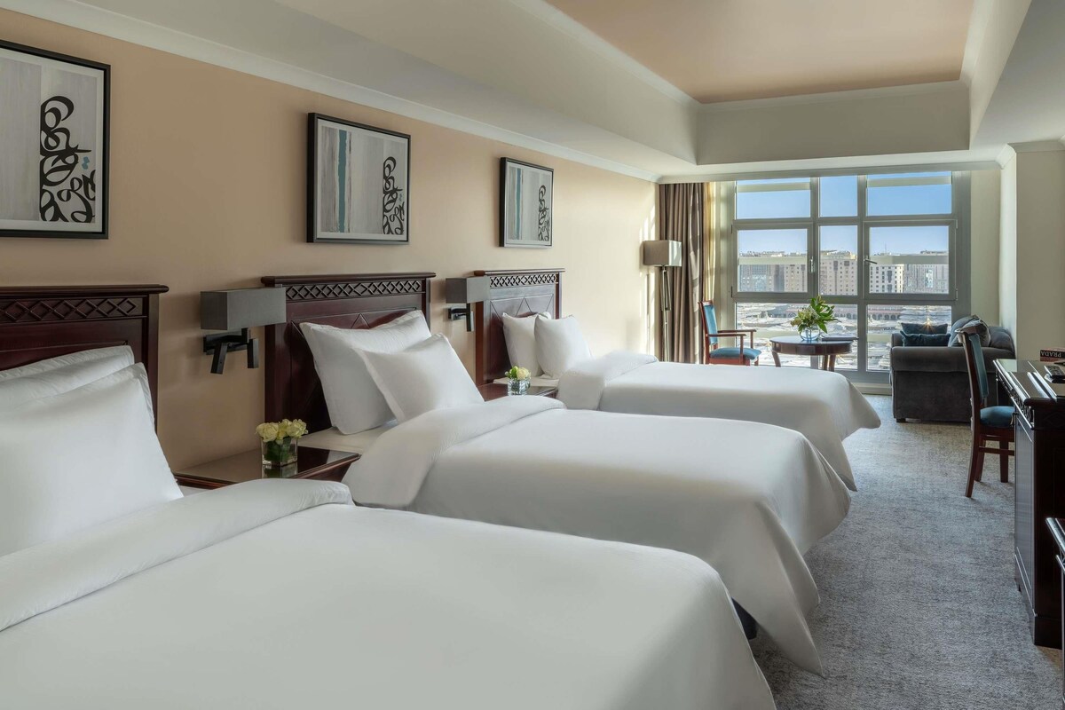 Habitación de hotel con tres camas, un escritorio, una silla y vistas a la ciudad.