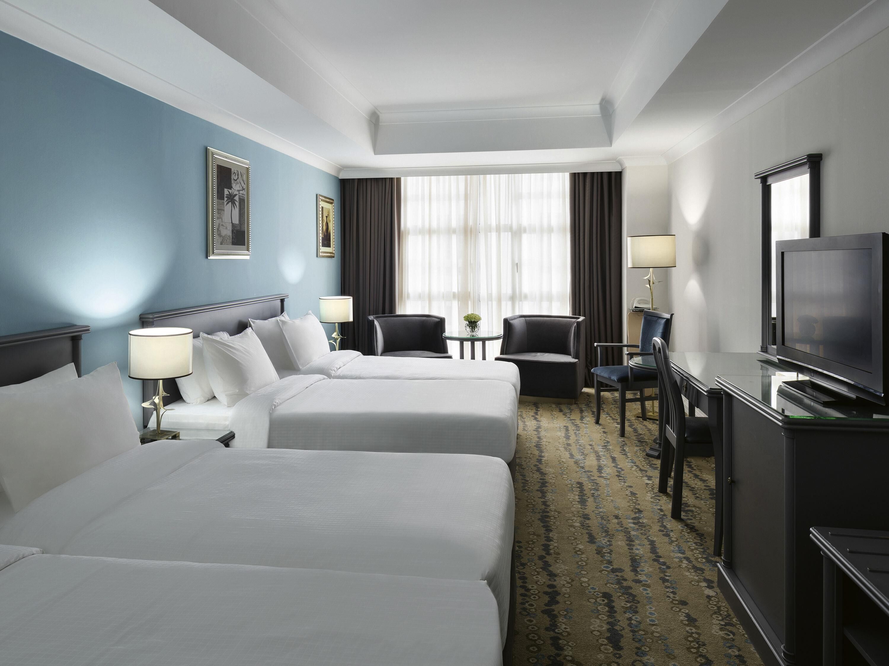 A hotel room with two beds, a desk with a television, a chair, and a small table with a lamp.
