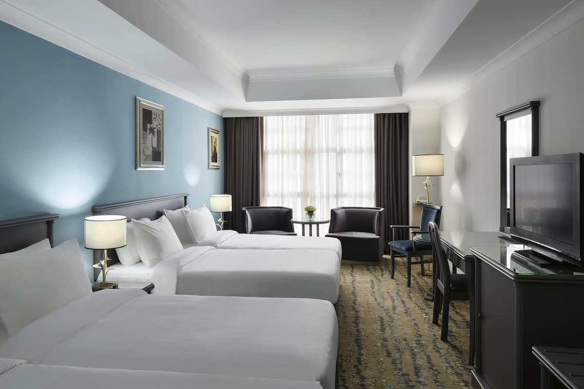 A hotel room with two beds, a desk with a television, a chair, and a small table with a lamp.