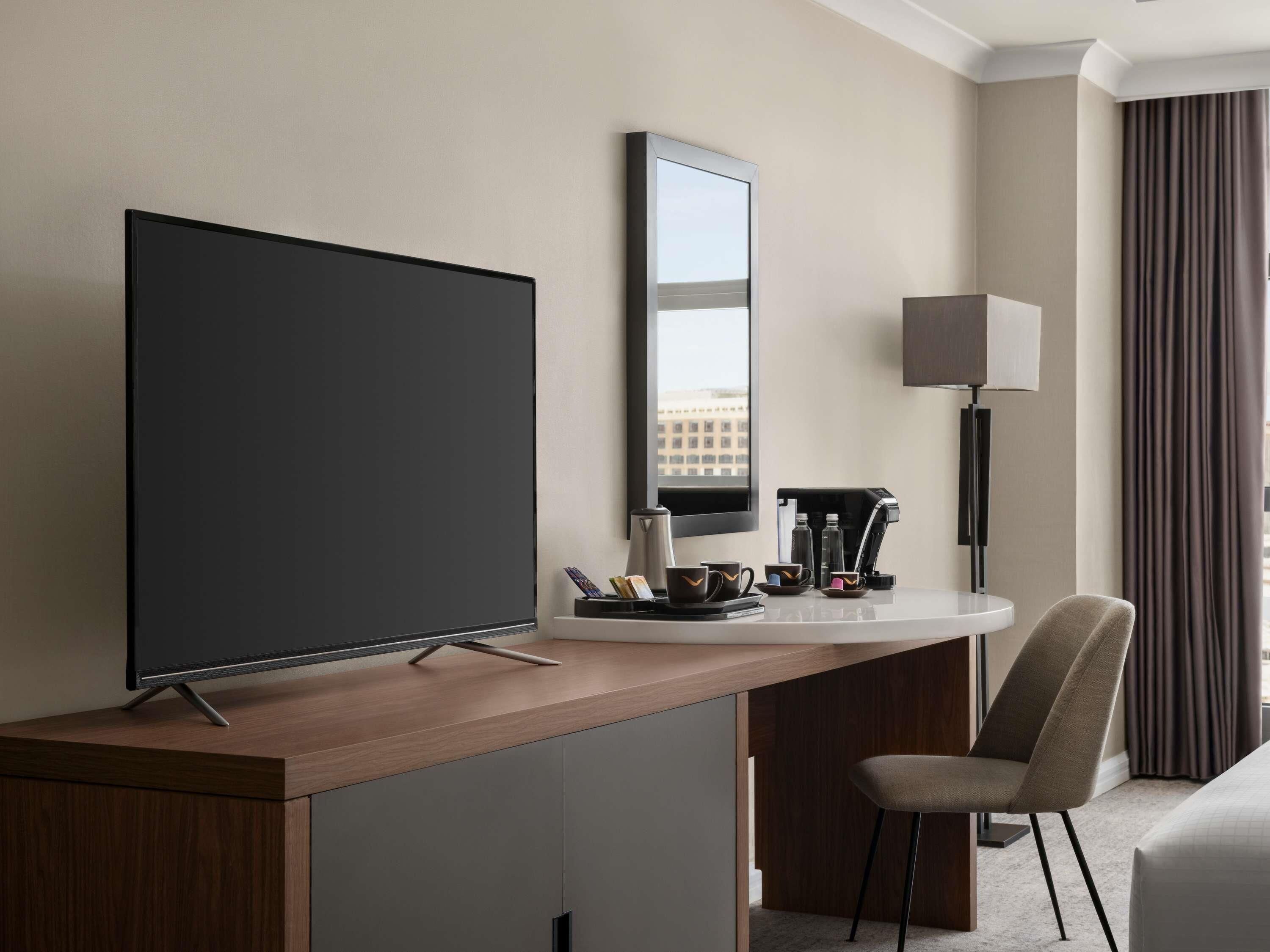 A hotel room with a large flat-screen TV, a coffee machine, a chair, and a bed.
