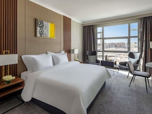 Quarto de hotel com uma cama grande, uma escrivaninha com abajur, uma cadeira, uma pequena mesa com jarra e vista para edifícios pela janela.