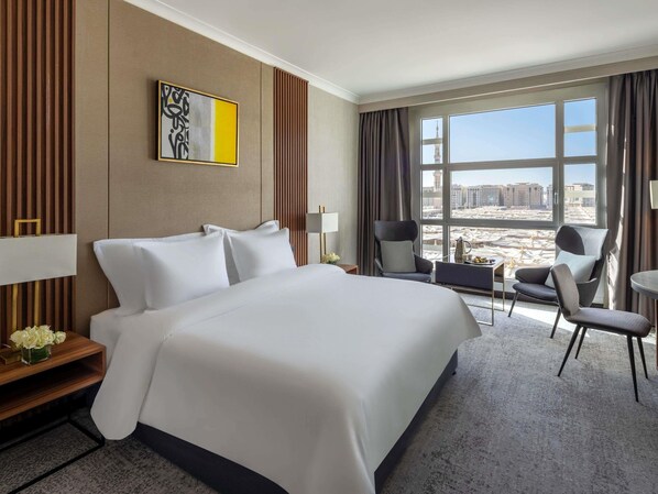 A hotel room with a large bed, a desk with a lamp, a chair, a small table with a pitcher, and a view of buildings through the window.