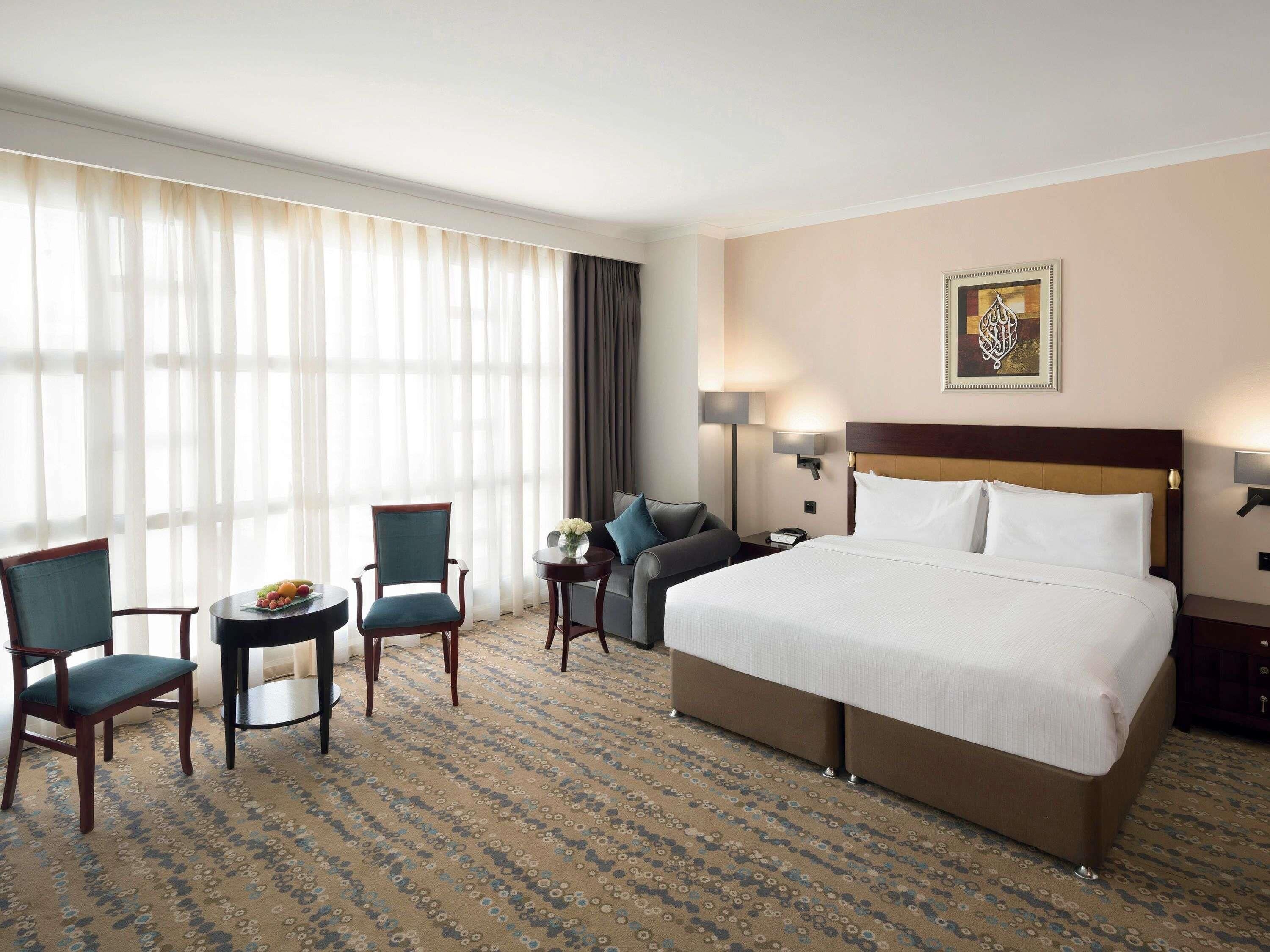 A hotel room with a large bed, two chairs, a small table with fruit, a lamp, and a painting on the wall.