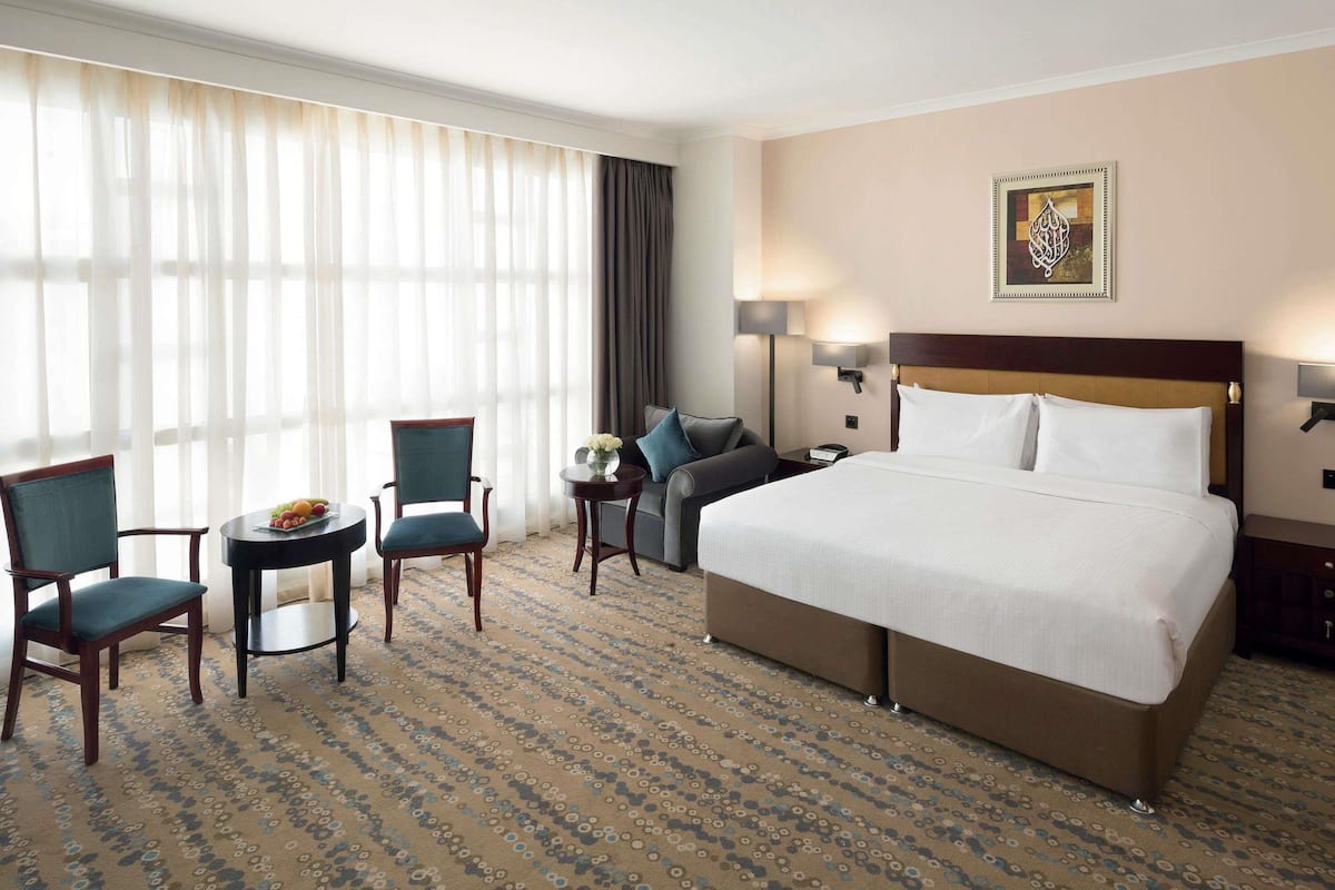 A hotel room with a large bed, two chairs, a small table with fruit, a lamp, and a painting on the wall.