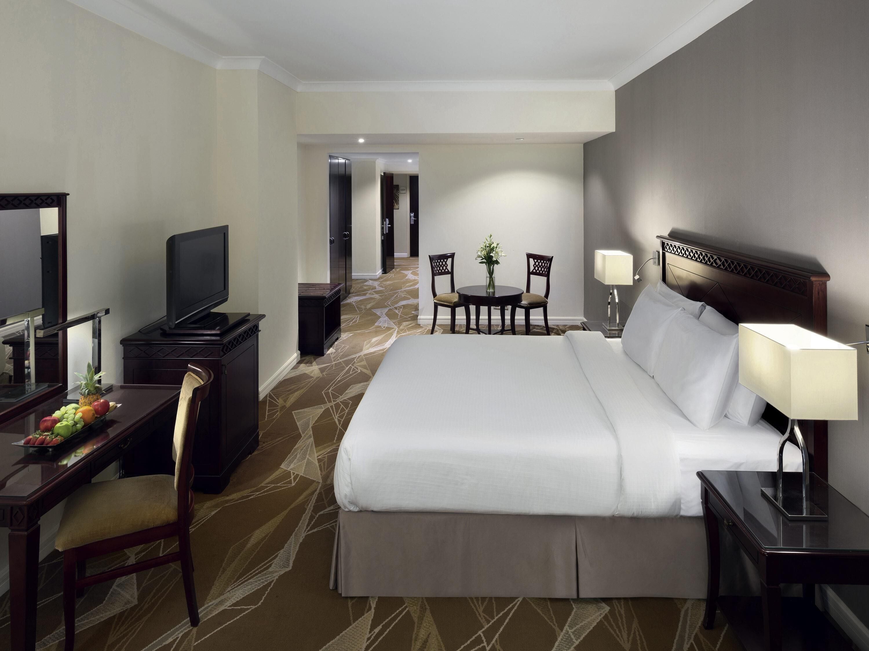 A hotel room with a bed, bedside table, lamp, TV, desk with fruit, and a chair.