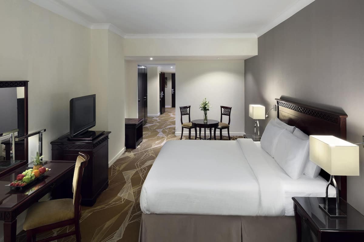 A hotel room with a bed, bedside table, lamp, TV, desk with fruit, and a chair.