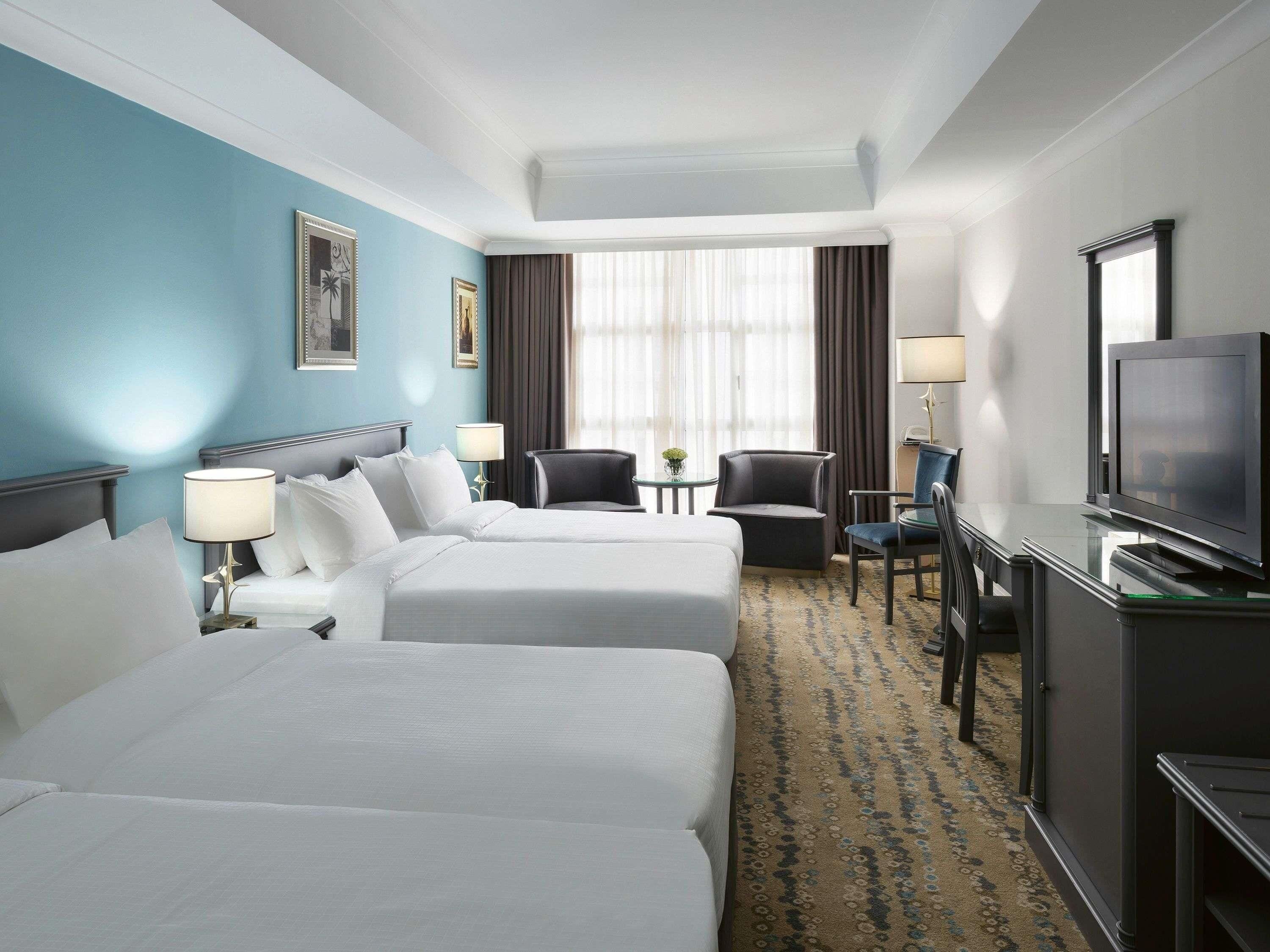 A hotel room with two beds, a desk with a television, a chair, and a small table with a lamp.
