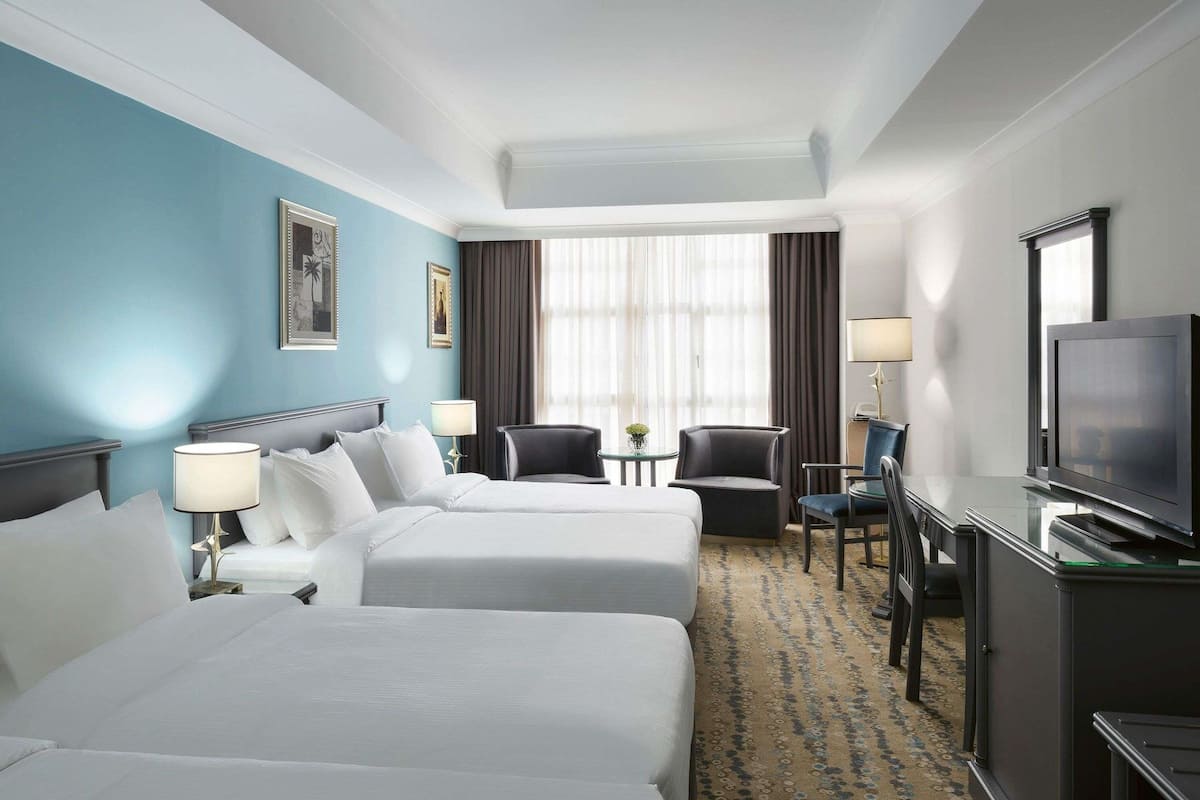 A hotel room with two beds, a desk with a television, a chair, and a small table with a lamp.