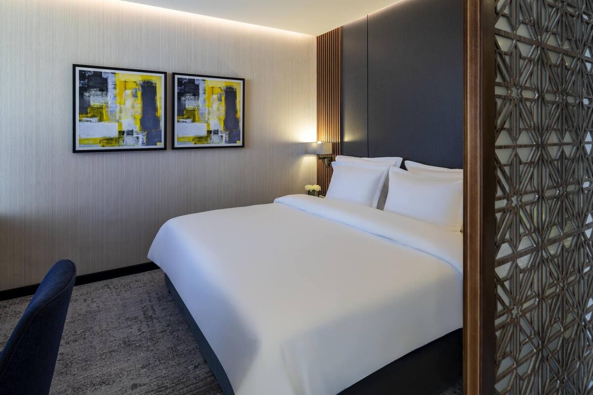 Hotellihuone, jossa on sänky, tuoli, kaksi abstraktia maalausta seinällä ja koristeellinen sermi.