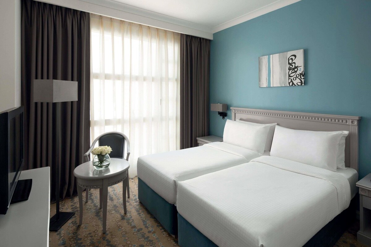 Habitación de hotel con dos camas, un escritorio, una silla, una mesita con un jarrón y una ventana con cortinas.