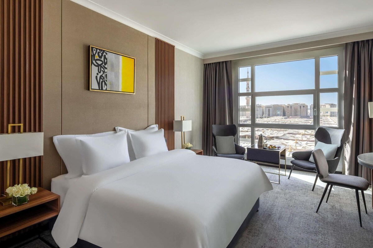 Una habitación de hotel con una cama grande, un escritorio con lámpara, una silla, una mesita con jarrón y vistas a edificios desde la ventana.