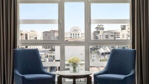 Une pièce avec deux chaises bleues, une petite table ronde et une vue sur le paysage urbain, visible à travers de grandes fenêtres.