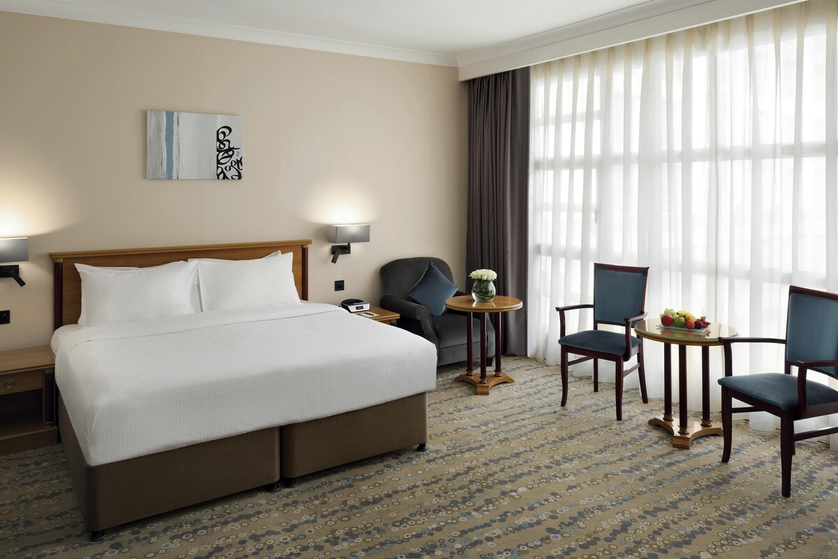 Habitación de hotel con una cama grande, dos sillones, una mesita con fruta, una lámpara y un cuadro en la pared.