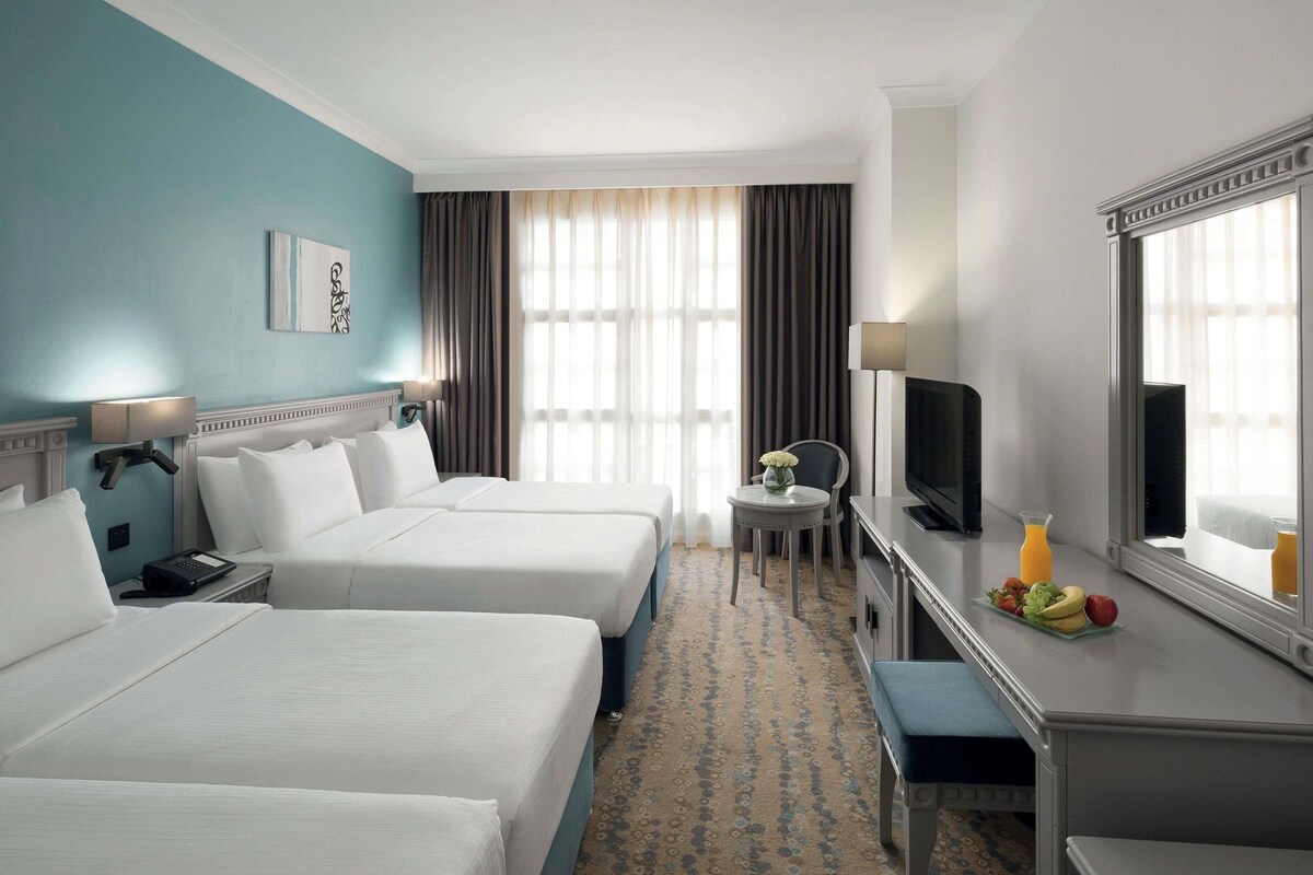 Habitación de hotel con dos camas, un escritorio con televisor, una silla, una mesita con fruta y un ventanal con cortinas.