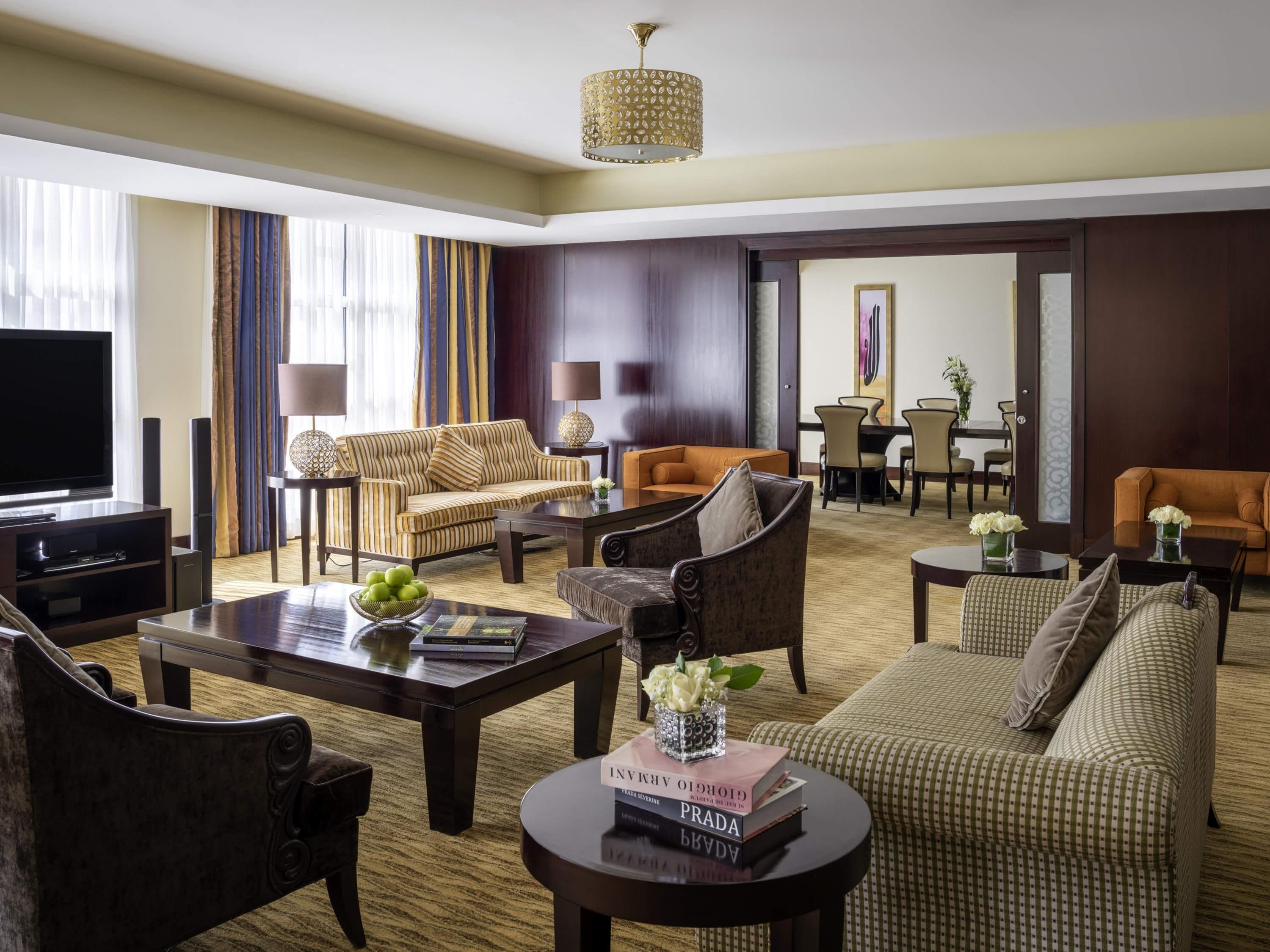 A hotel living room with a television, sofas, a dining area, and a chandelier.