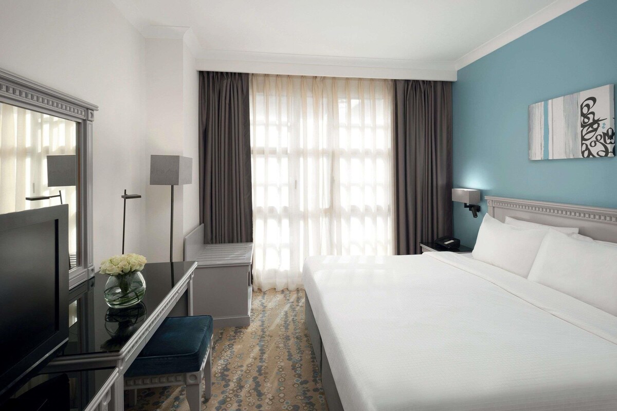 Habitación de hotel con cama, escritorio, silla, televisión, lámpara y ventana con cortinas.