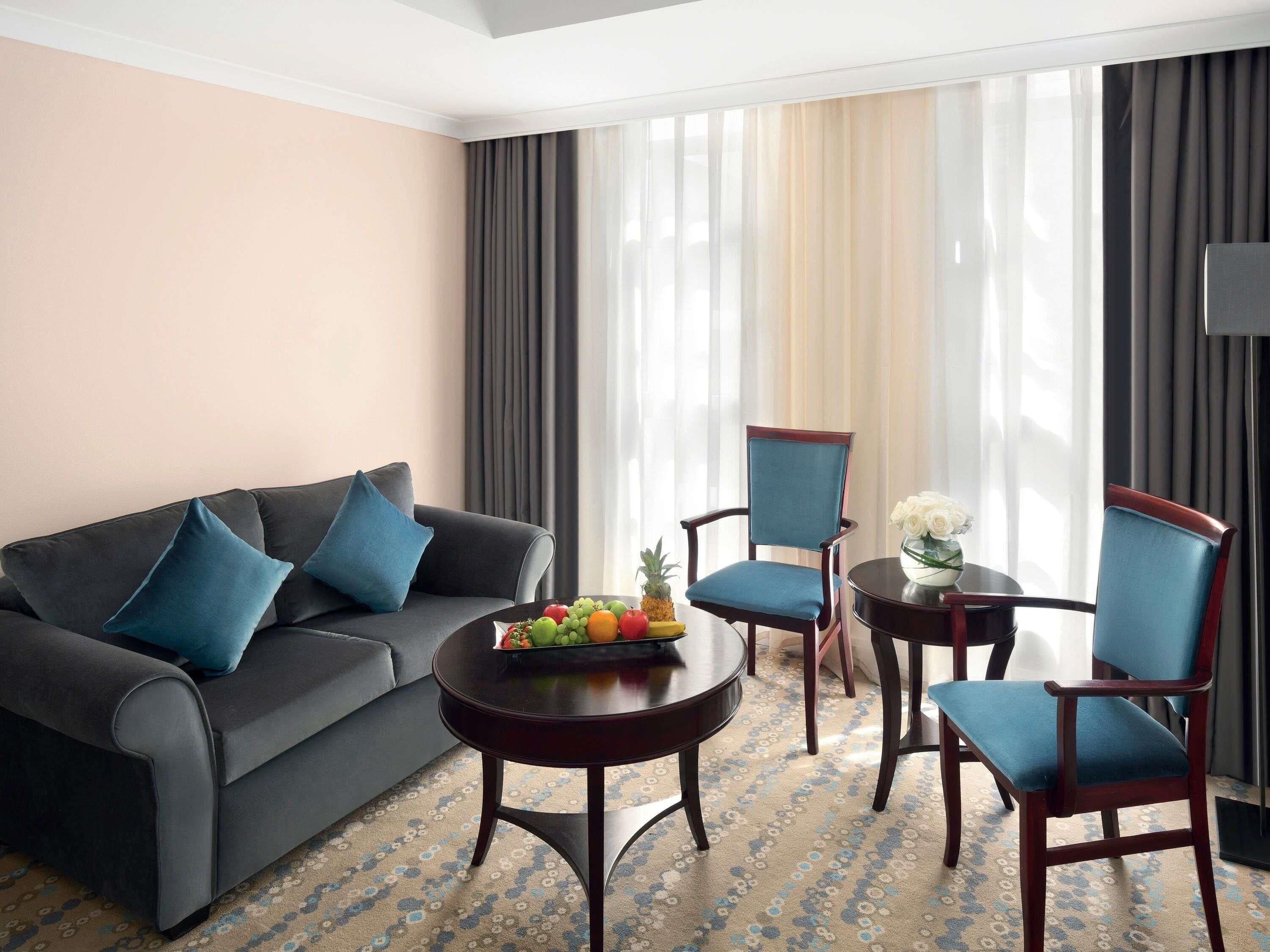 Una habitación de hotel con un sofá gris oscuro, dos sillones azules, una mesa de centro redonda de madera con fruta y un jarrón de flores blancas.