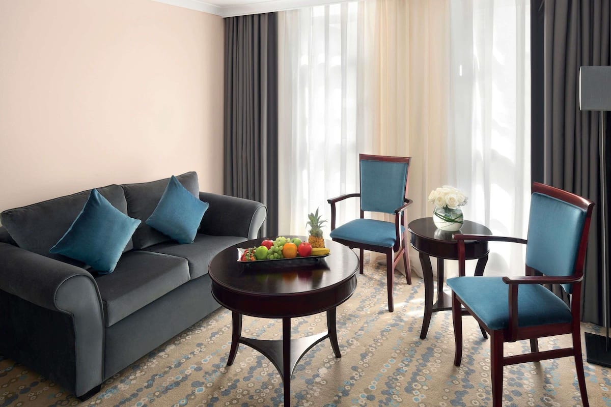 Una habitación de hotel con un sofá gris oscuro, dos sillones azules, una mesa de centro redonda de madera con fruta y un jarrón de flores blancas.