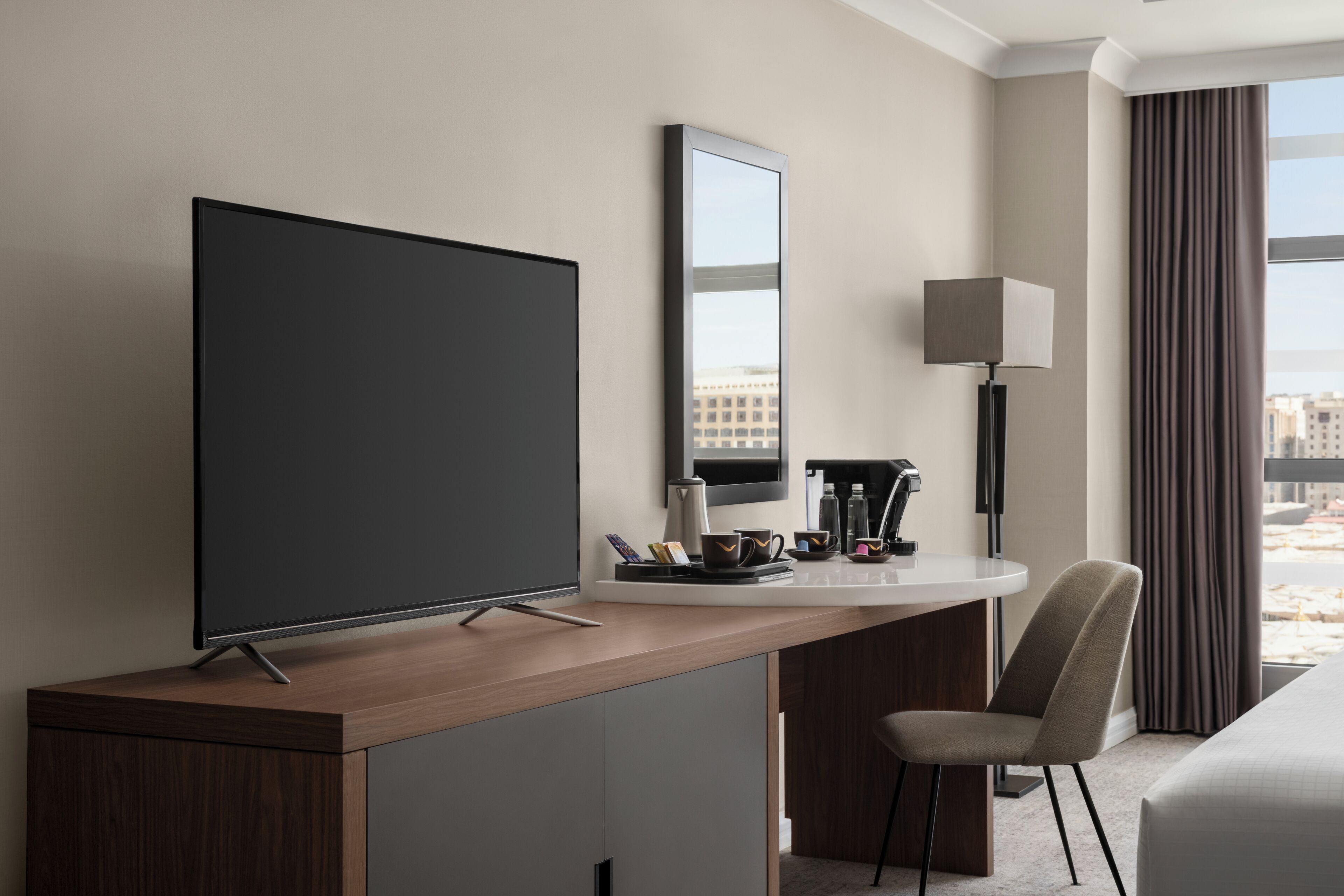 Una habitación de hotel moderna con una televisión de pantalla plana, un escritorio con lámpara, una silla y un amplio ventanal con cortinas.