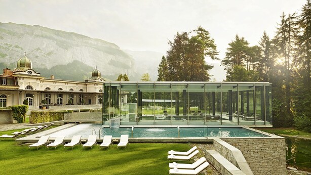 A modern glass-enclosed pool area with lounge chairs, surrounded by a well-manicured lawn and a historic building with domed roofs. - Chalet Belmont Hotel (Flims)