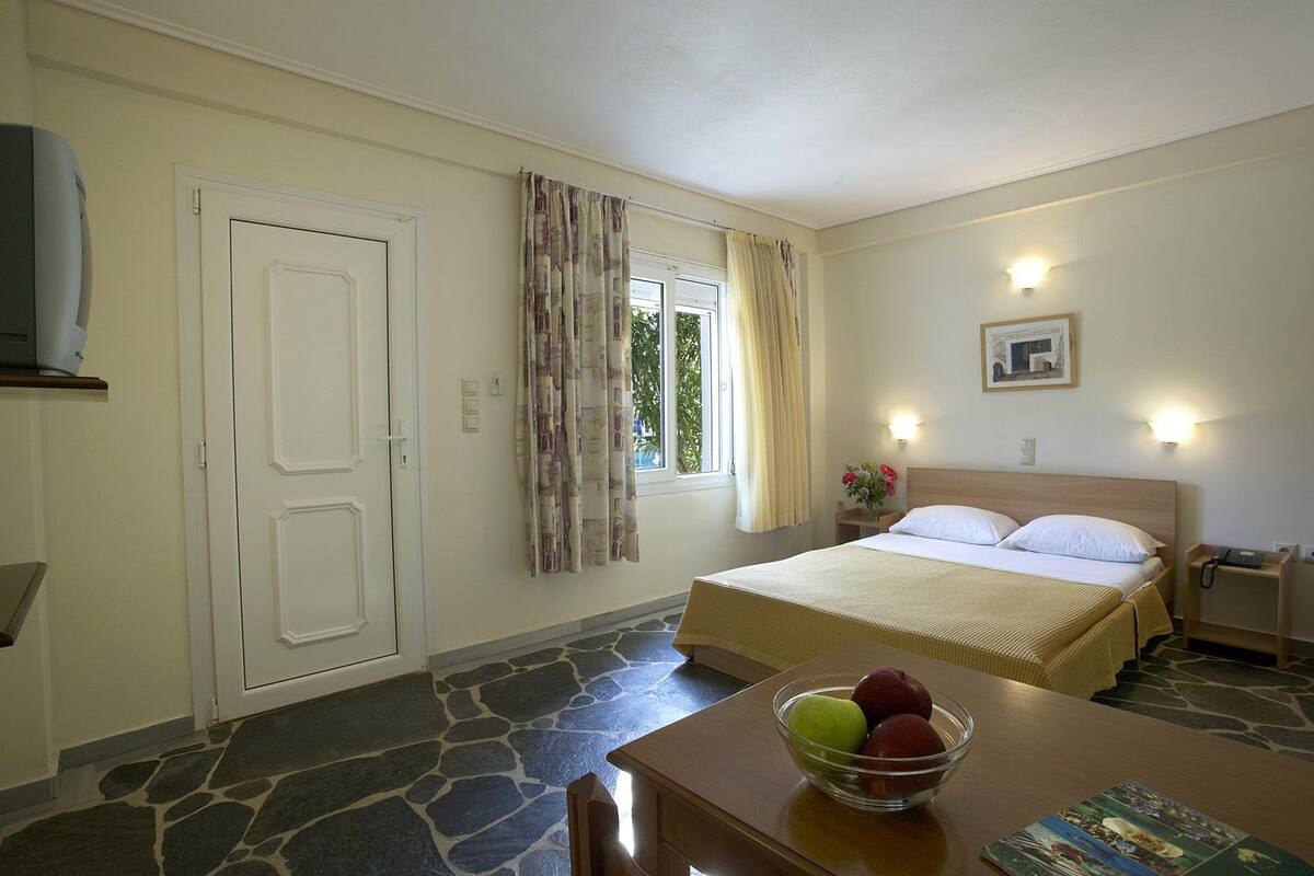 A bedroom with a bed, a window with curtains, a bedside table with a lamp, and a bowl of fruit on the table.