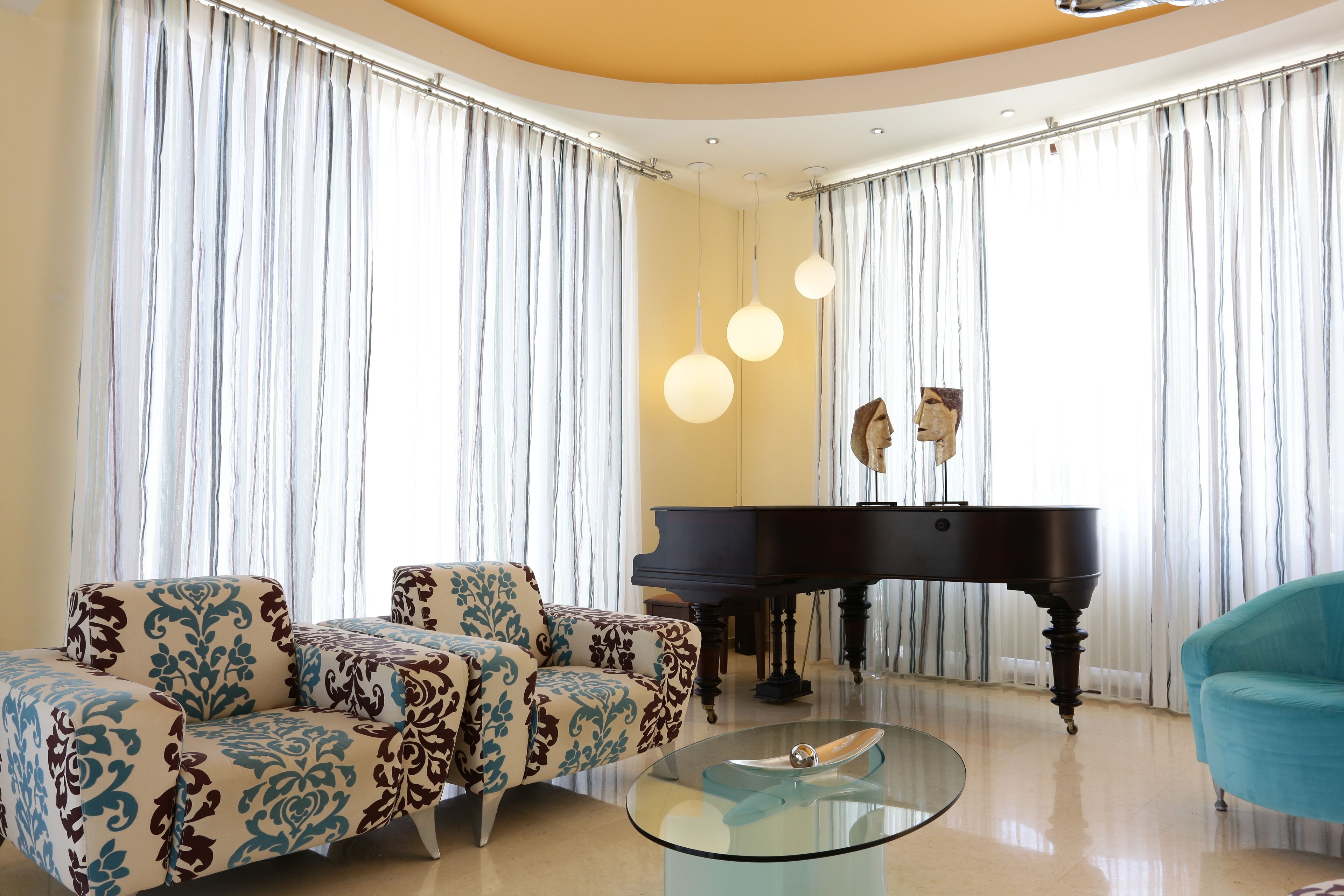 A living room with a grand piano, patterned armchairs, a glass coffee table, and a teal armchair.