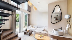 A modern living room with a staircase, a sofa, a coffee table, and a bathroom with a round sink and mirror. - Alexandra Golden Boutique Hotel (Thasos)
