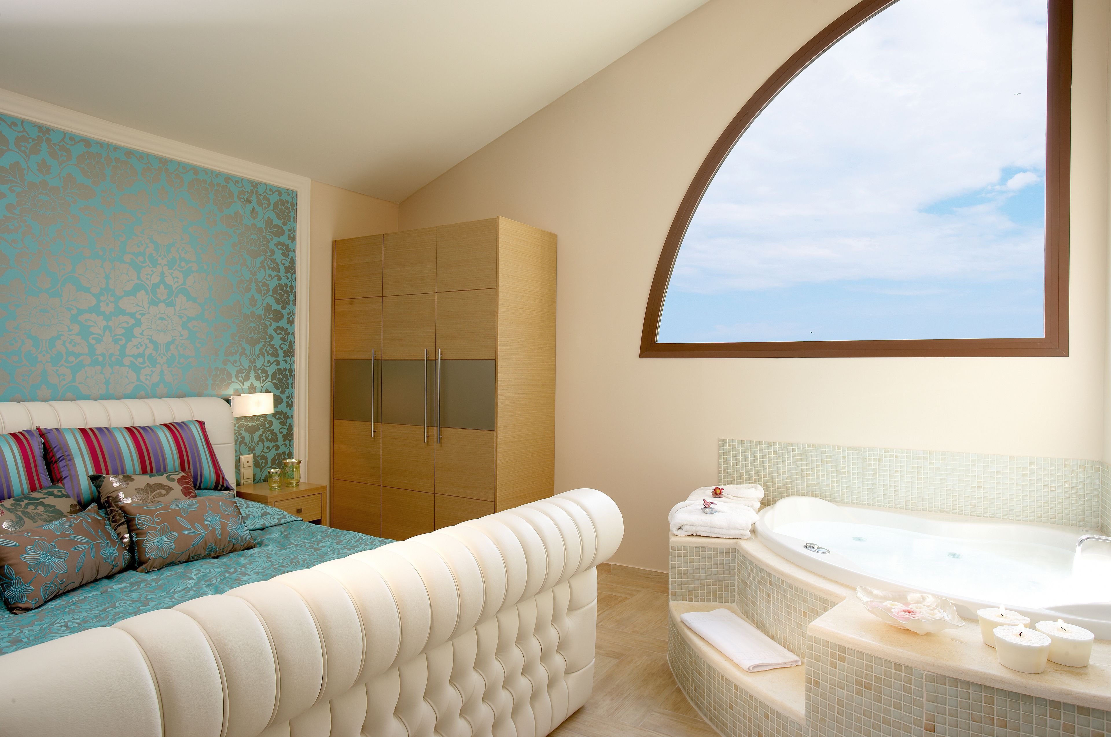 Une chambre avec un grand lit, une armoire en bois, une salle de bain avec une baignoire et une vue sur le ciel par une fenêtre.