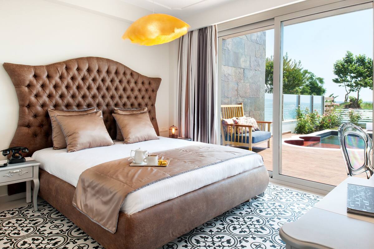 Een slaapkamer met een groot, gestoffeerd hoofdeinde, een bed met een deken in wit en bruin, een nachtkastje met een lamp, en uitzicht op een buitengebied met een zwembad en groen.