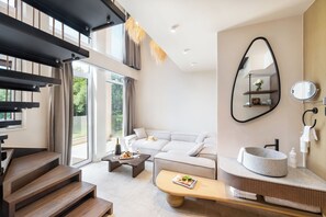 A modern living room with a staircase, a sofa, a coffee table, and a bathroom with a round sink and mirror. - Alexandra Golden Boutique Hotel (Thasos)