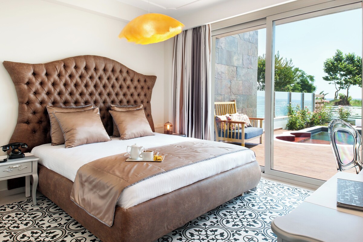 Een slaapkamer met een groot, gestoffeerd hoofdeinde, een bed met een deken in wit en bruin, een nachtkastje met een lamp, en uitzicht op een buitengebied met een zwembad en groen.