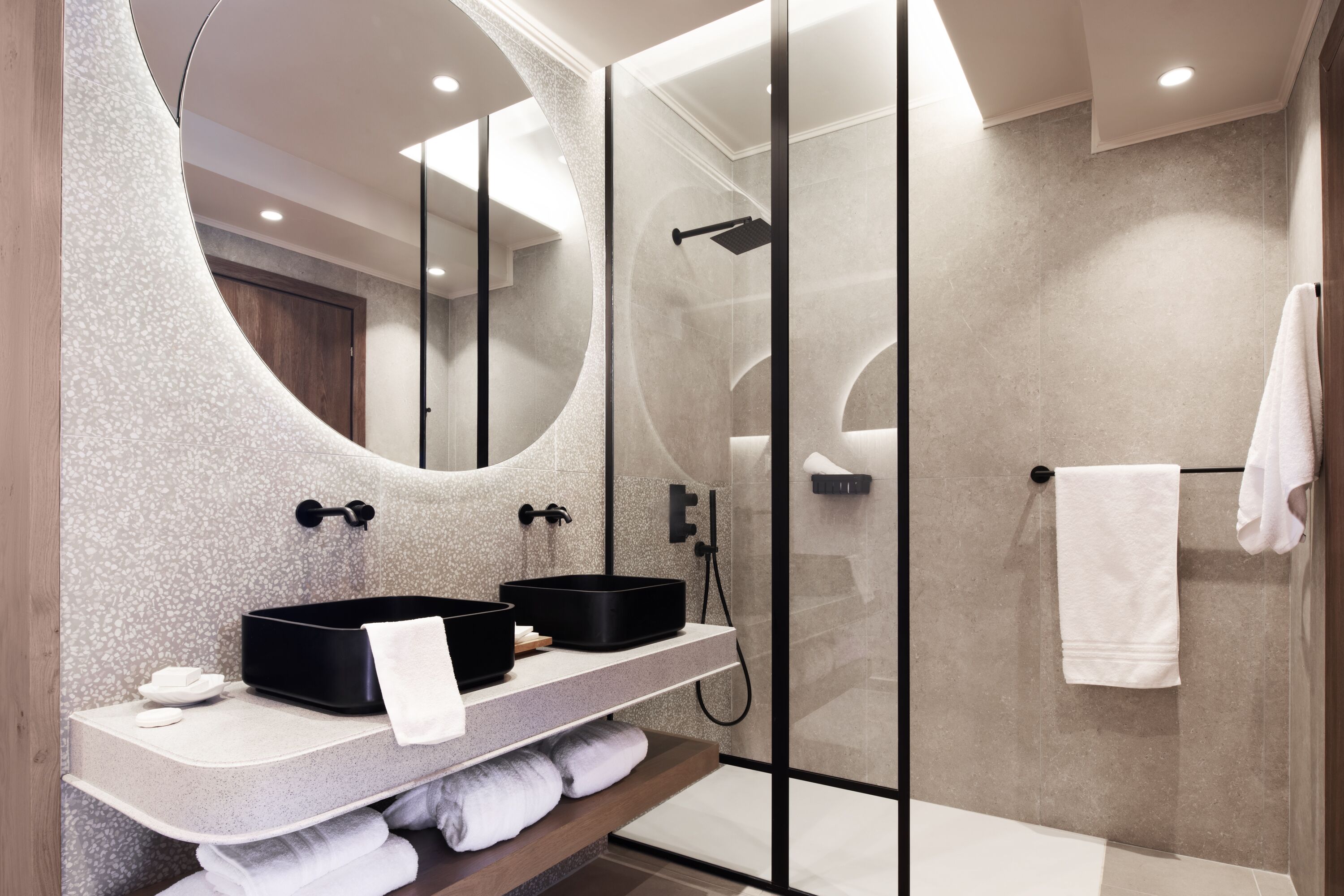 Une salle de bain moderne avec un miroir incurvé, deux vasques noires, une douche en verre et une serviette blanche suspendue à un porte-serviettes.