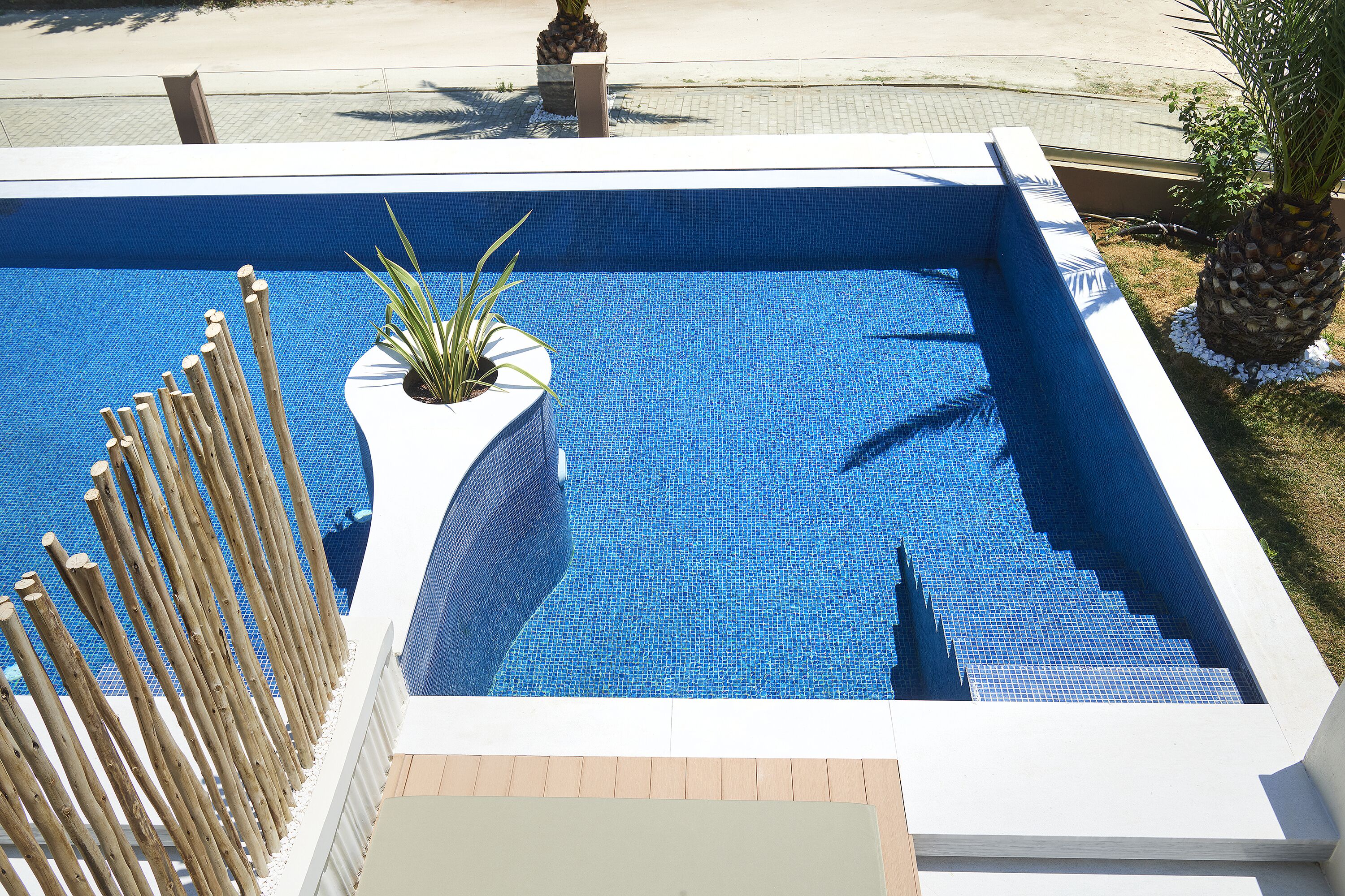 Une piscine rectangulaire aux carreaux de mosaïque bleus, entourée d’une terrasse blanche et d’une clôture en bambou.