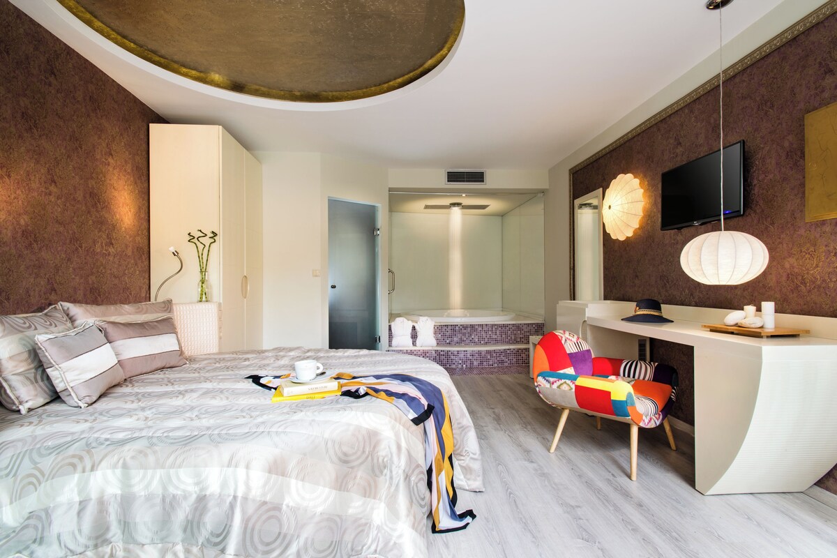Een moderne hotelkamer met een bed, een bureau, een televisie, een badkamer en een kleurrijke stoel.