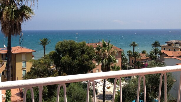 A balcony with a view of the sea, palm trees, and buildings with red-tiled roofs. - Hotel Nettuno (Diano Marina)