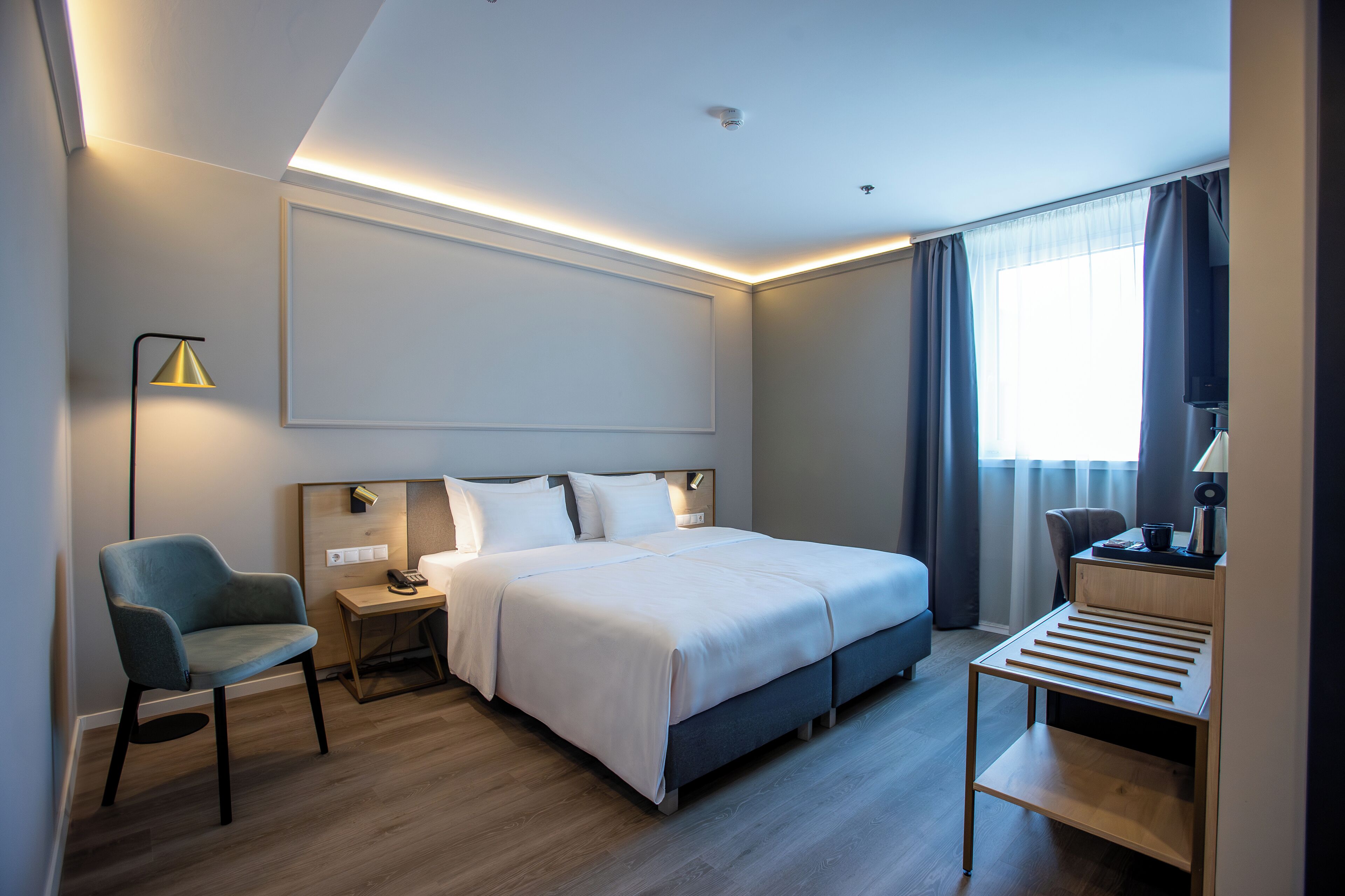 A modern hotel room with a neatly made bed, a desk with a lamp, a chair, and a large window with curtains.