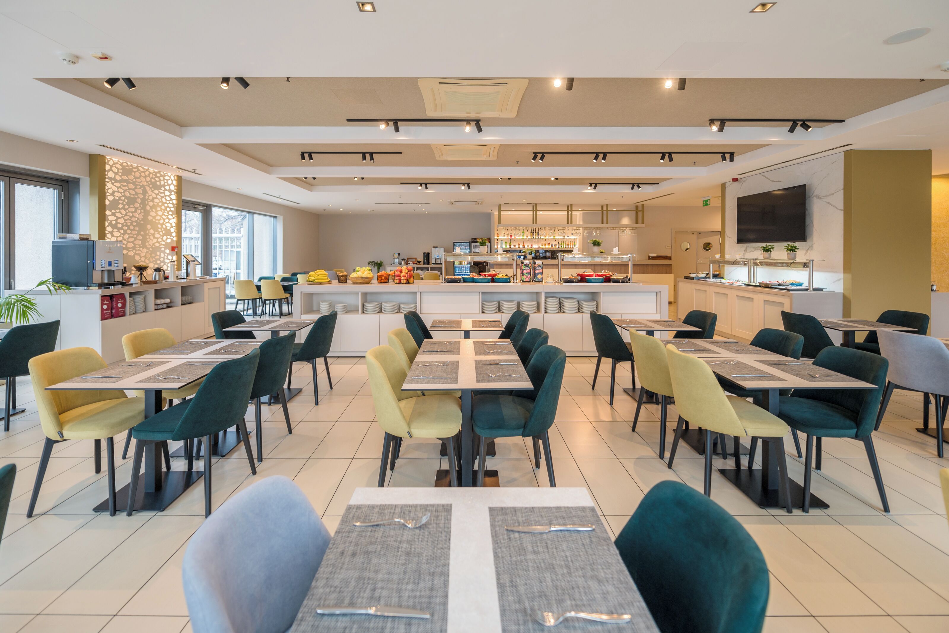 Un espace repas moderne, doté de plusieurs tables et chaises, d’un comptoir central où sont exposés les plats, et d’un espace cuisine en arrière-plan.