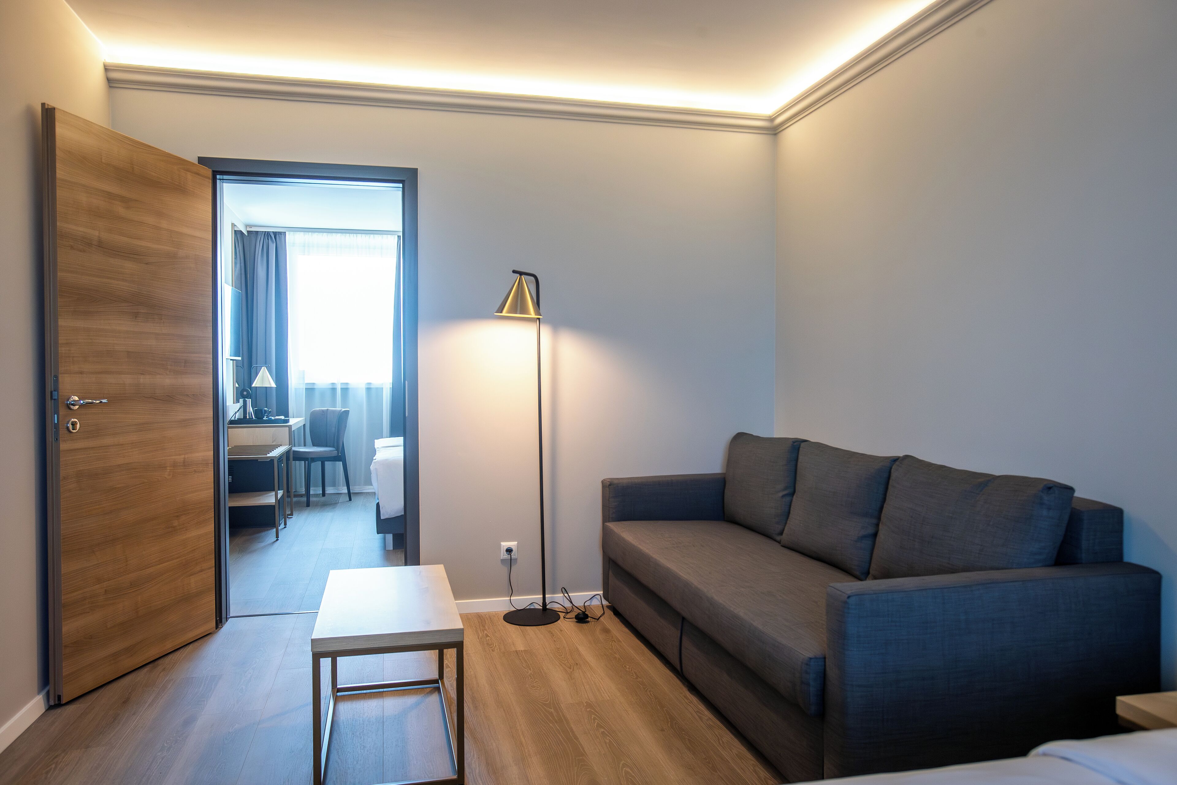 Une chambre d’hôtel moderne avec une porte en bois, un canapé gris, une petite table et un lampadaire.