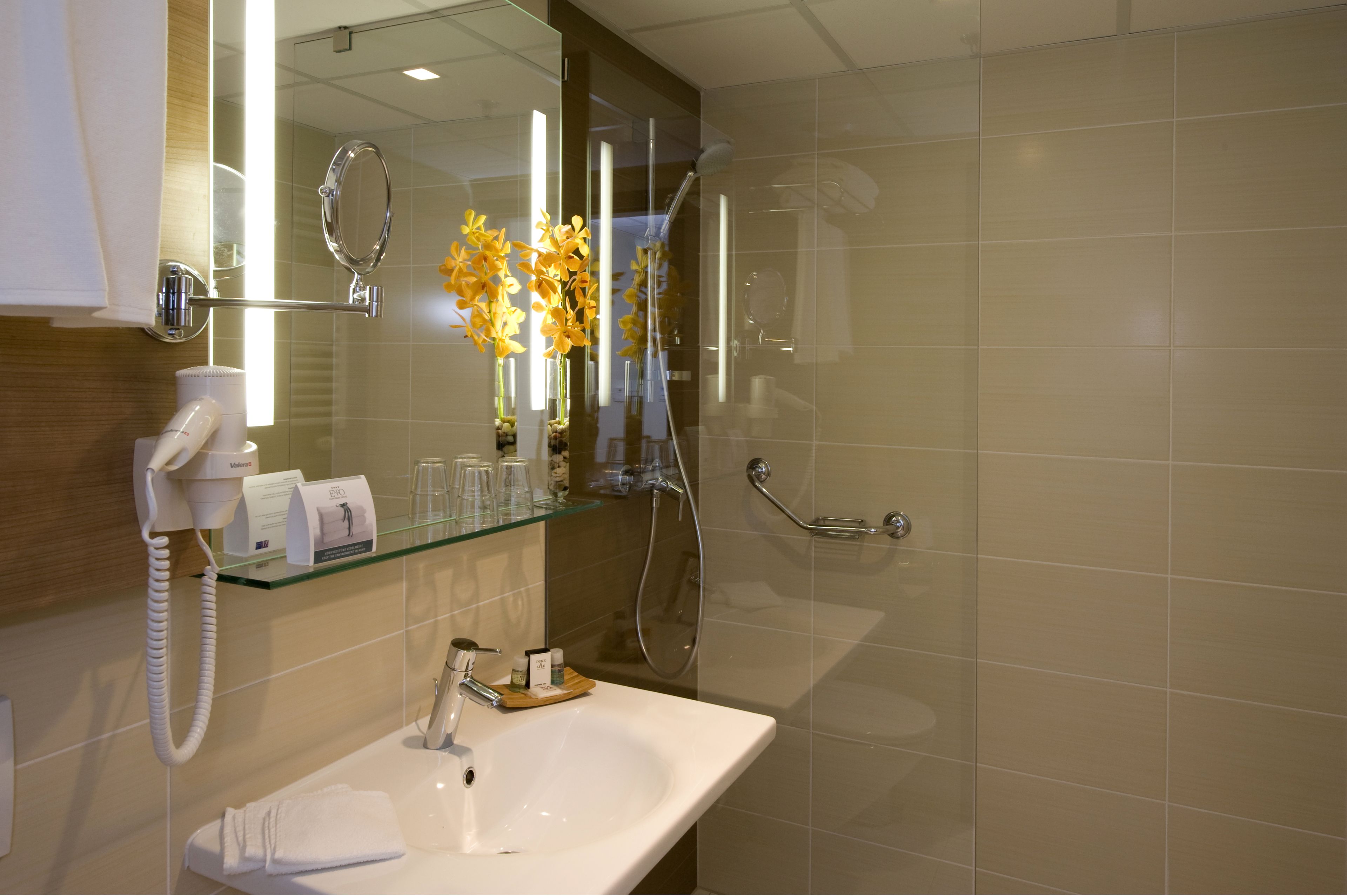 A modern bathroom with a glass shelf holding toiletries, a white sink, a hairdryer, and a mirror with a ring light.