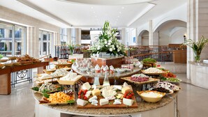 A buffet spread with various cheeses, meats, and salads in a spacious hotel dining area with large windows and a high ceiling. - Golden Crown Hotel (Nazareth)
