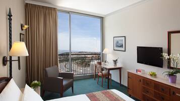 A hotel room with a bed, a desk, a chair, a television, and a view of the city through large windows.