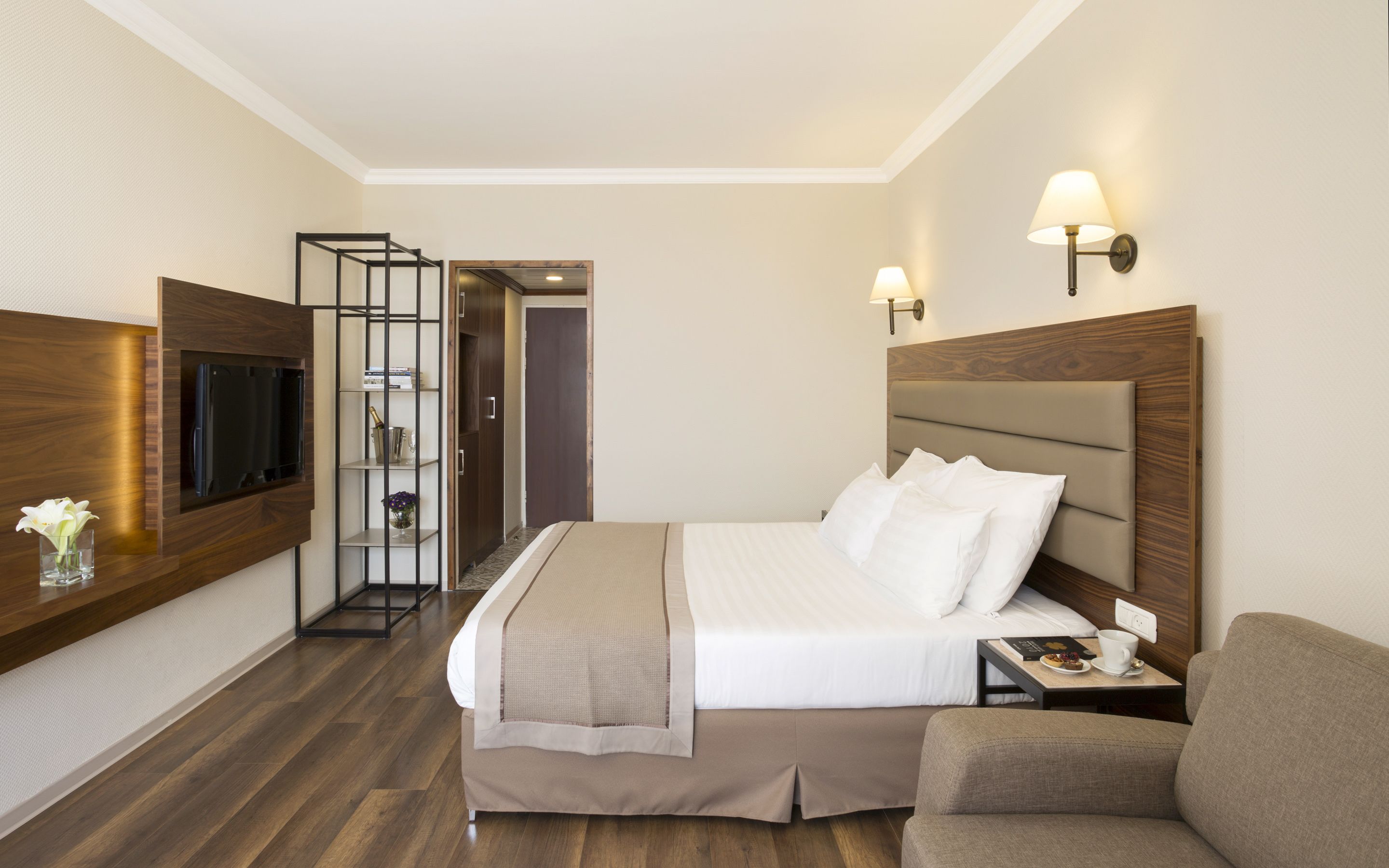 A hotel room with a bed, a TV mounted on the wall, a bedside table with a lamp, a chair, and a small table with a cup and a book.